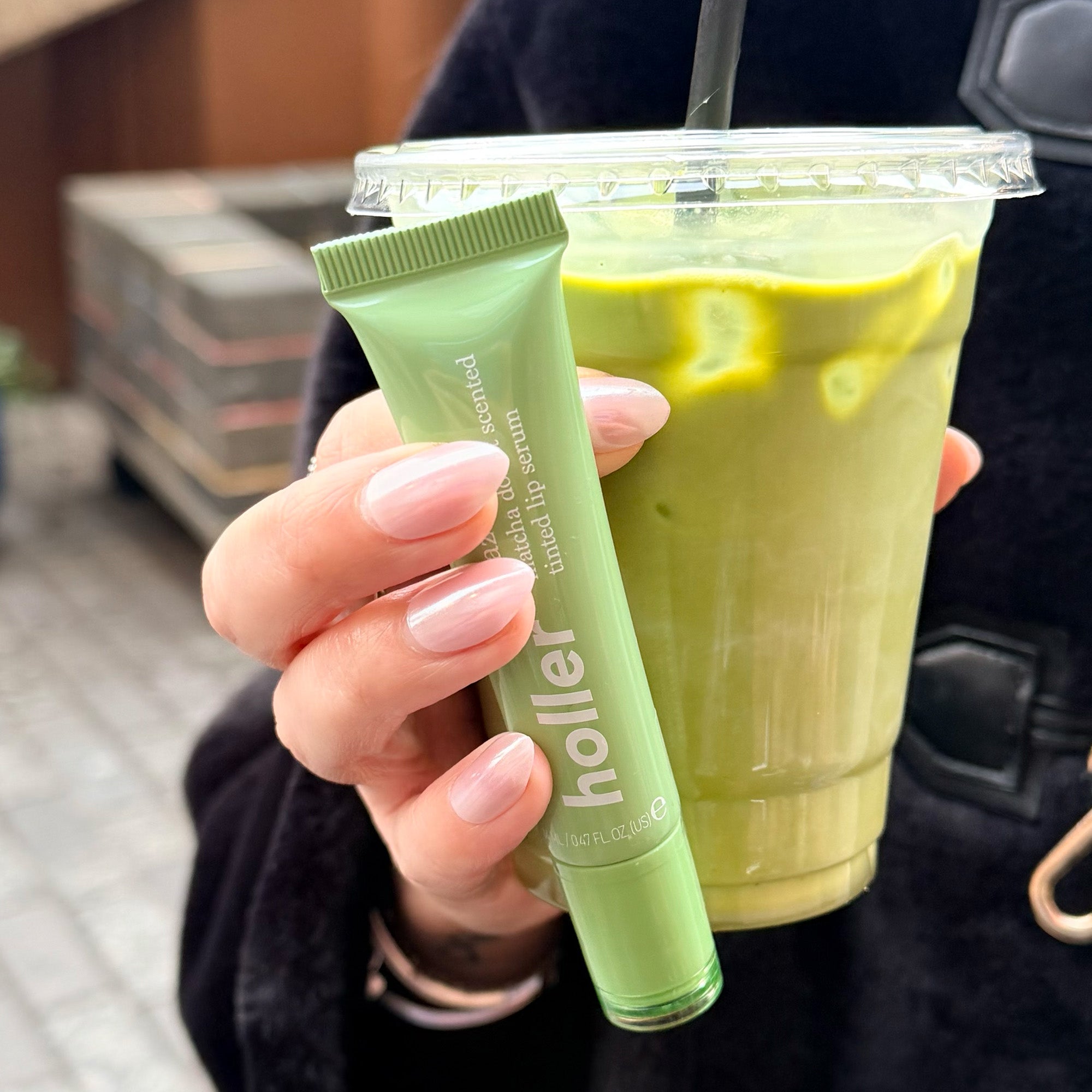 Girl holding a Lip gloss with "Holler" on the tube and a green matcha against a black jacket. 