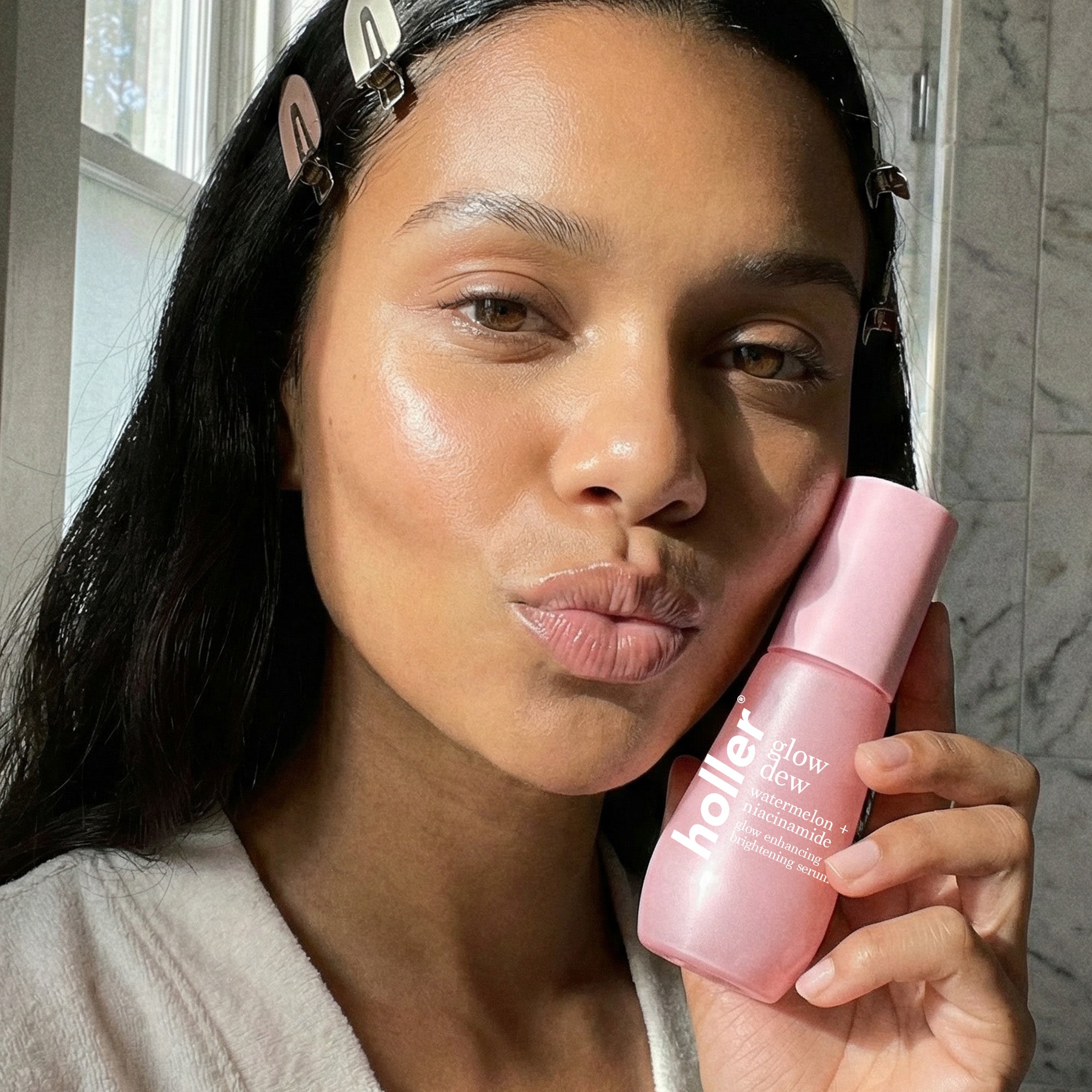 Woman holding a pink bottle of 'Holler glow dew' product in a bathroom setting